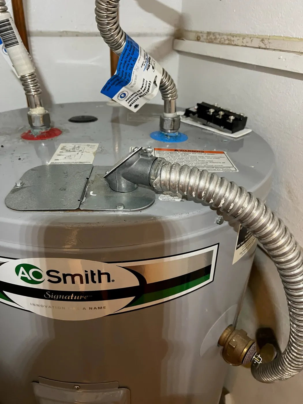 The top of an installed A. O. Smith Signature electric water heater showing flexible water connectors and a flexible electrical conduit run into the junction box.